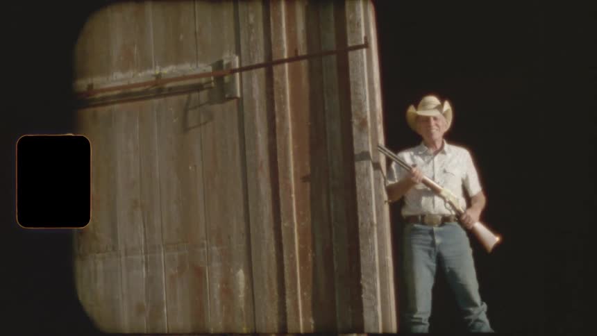 An old cowboy in a hat proudly holds a rifle in the doorway of a rustic barn, captured on vintage film
