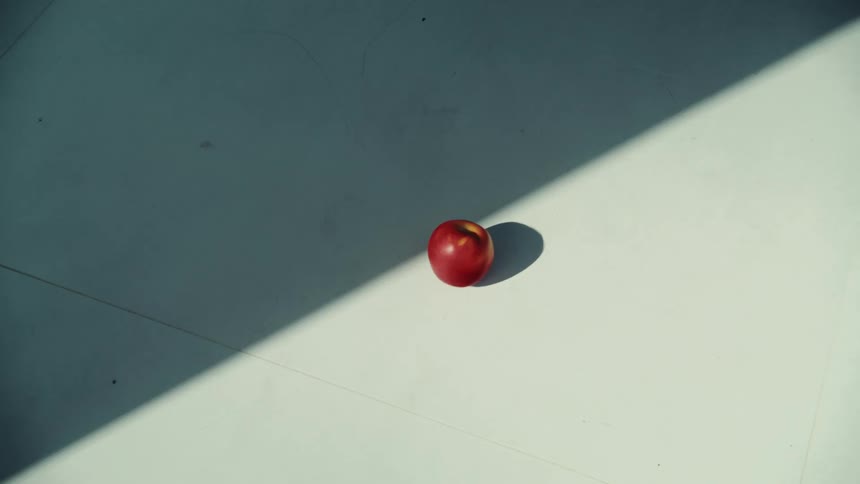 A single red apple spins and then slowly rolls from a beam of light into the shadows on a white floor