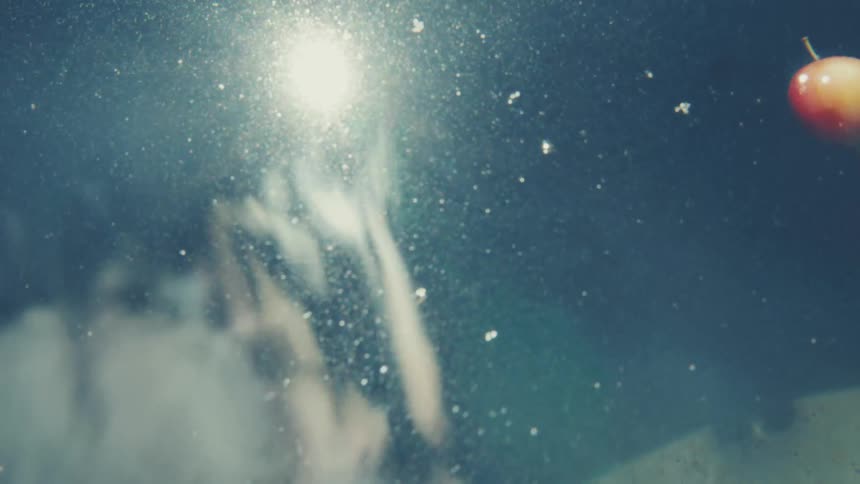 Dreamy underwater view looking up at a sunlit surface as an apple falls into the dark, rippling water