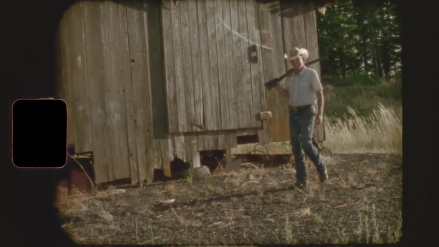 Vintage 8mm footage shows a cowboy with a rifle walking slowly past a weathered wooden outbuilding in a rural setting