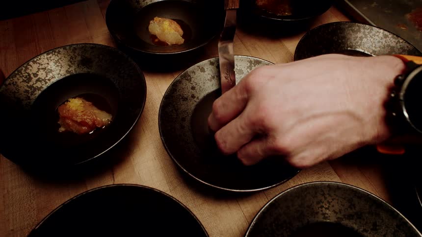 A chef meticulously plates a gourmet raw fish appetizer into several small, rustic bowls on a wooden counter