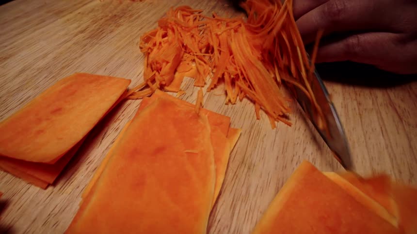 Close-up of a person's hands finely chopping fresh carrots into thin strips on a wooden cutting board