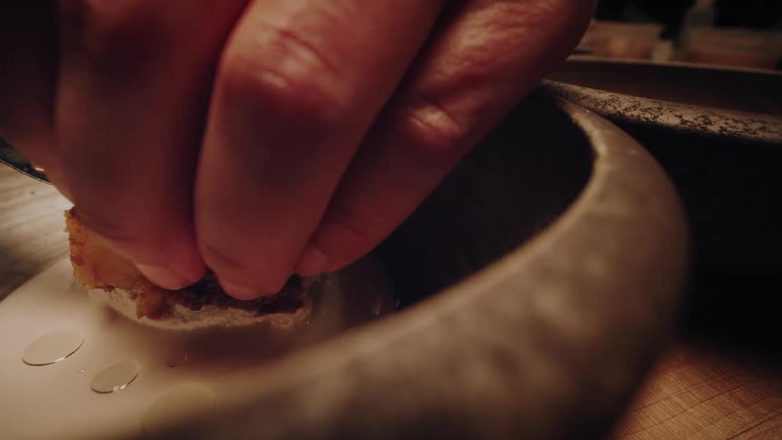 A chef's hand carefully places a seasoned piece of food onto a creamy base inside a rustic bowl, preparing a gourmet dish