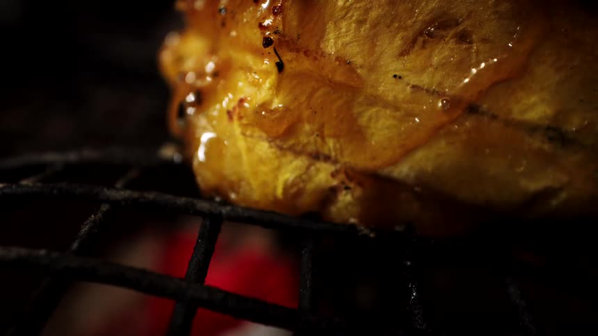 Extreme close-up of a plantain being glazed with a pink brush while grilling over glowing red embers