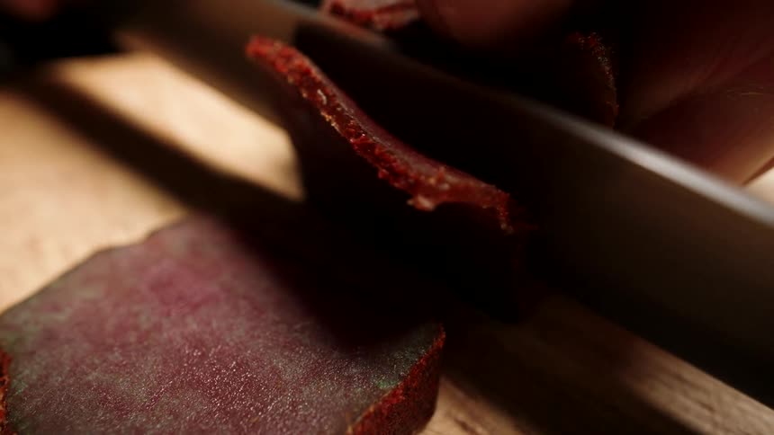 A macro view captures a chef's hands thinly slicing spiced, cured meat with a sharp knife on a wooden cutting board