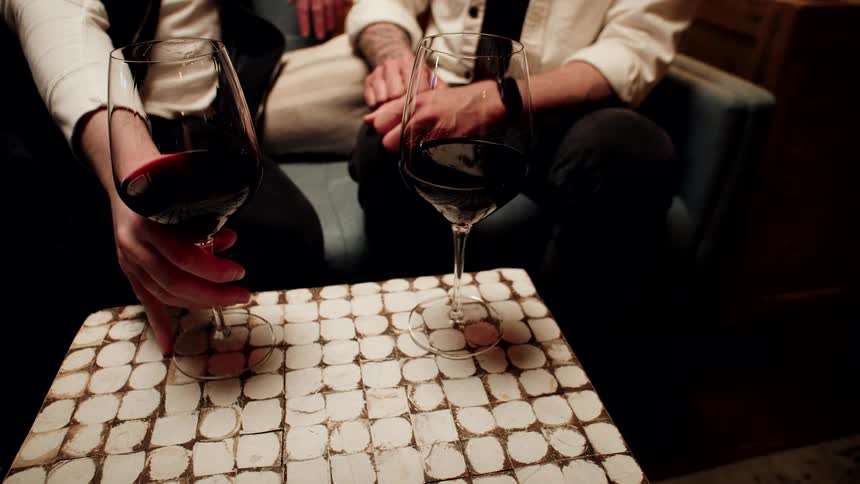 A man swirls a glass of red wine in a dimly lit lounge while sitting with another person