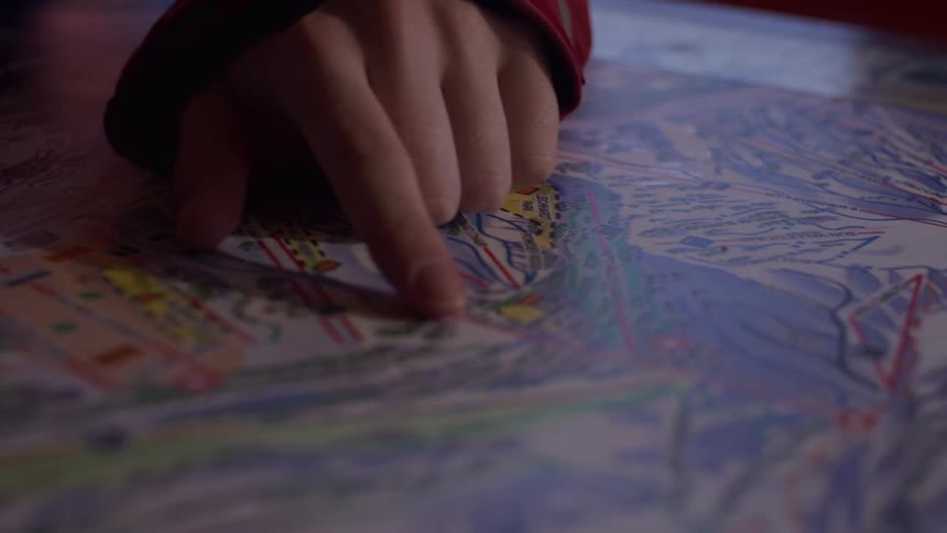 A young skier in a red jacket and helmet plans their route by tracing a finger across a ski resort trail map