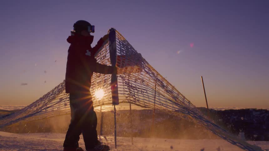 A ski patroller, silhouetted against the rising sun, shakes snow off a safety net in dramatic slow motion on a mountaintop