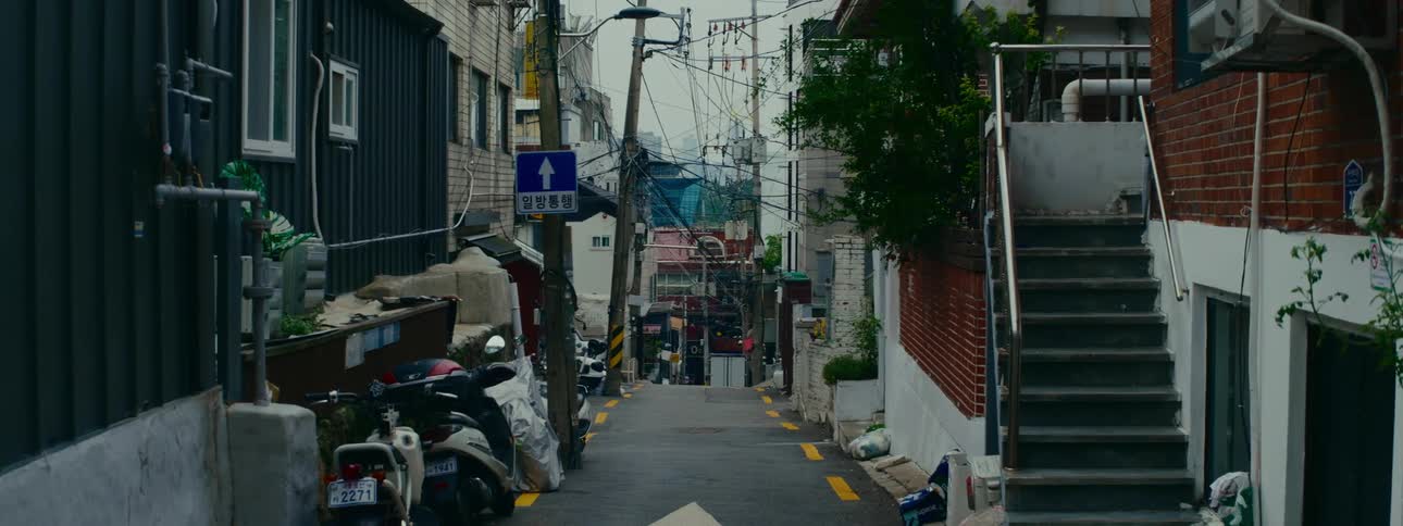 A static view down a steep, narrow alley in Seoul, South Korea, with a complex web of overhead power lines and parked scooters