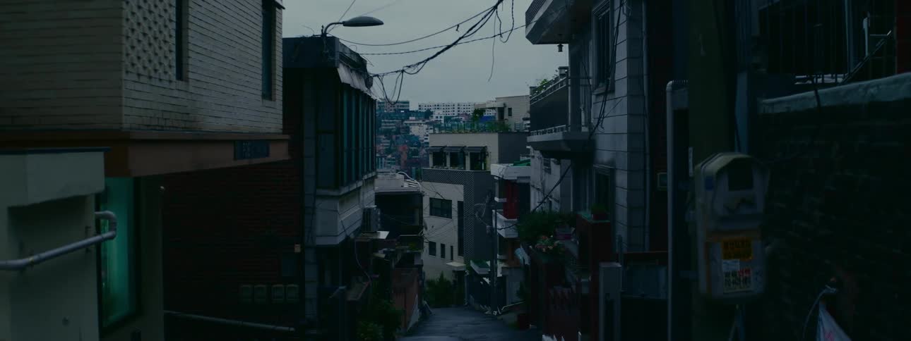 A moody, static shot looks down a steep and narrow alleyway in a dense residential neighborhood of a city on an overcast day