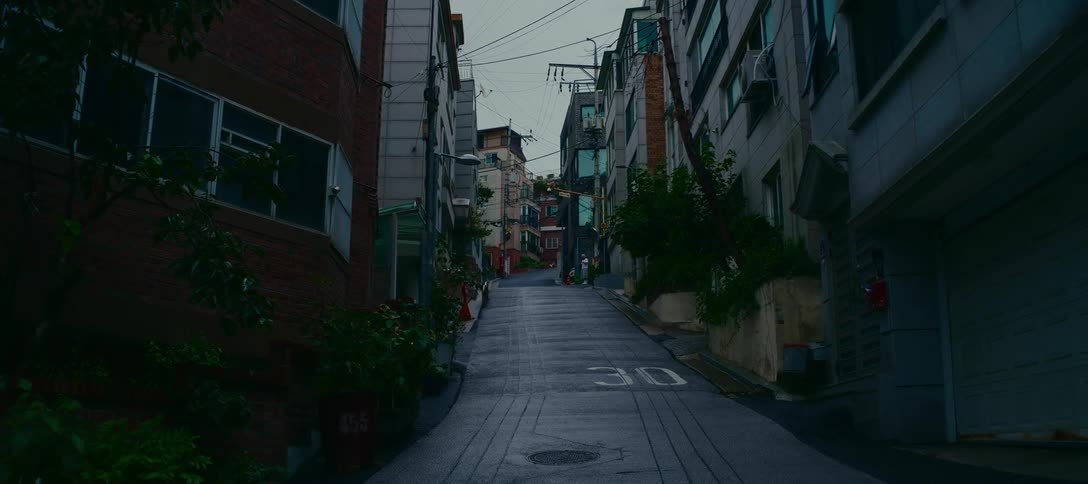 A moody static shot of a steep, empty street winding uphill through a quiet residential neighborhood on an overcast day