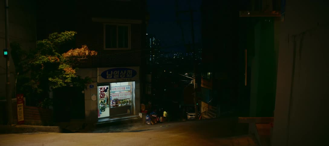 A quiet, empty street corner in a South Korean neighborhood at night, with a brightly lit shop and the city skyline in the background