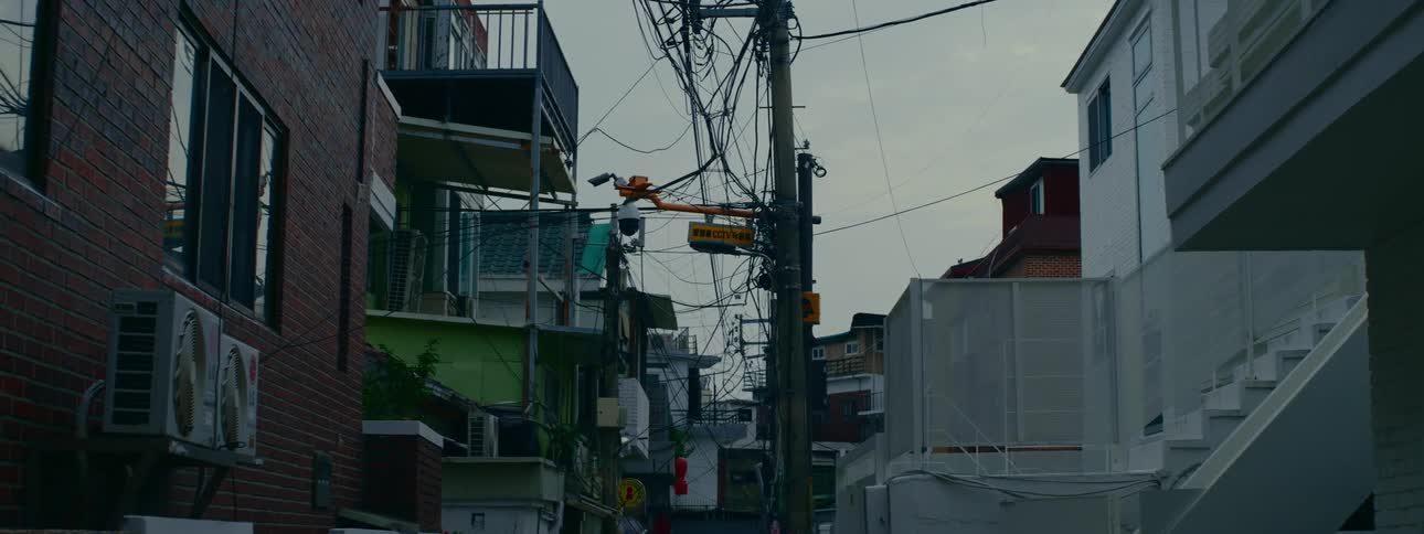 A chaotic tangle of electrical wires hangs from a utility pole in a narrow residential alley in South Korea under a moody, overcast sky