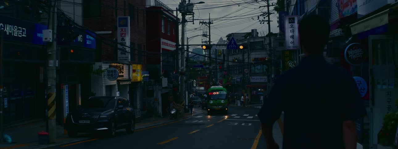 A green bus with a cartoon face drives down a busy, narrow street in Seoul, South Korea during a gloomy evening
