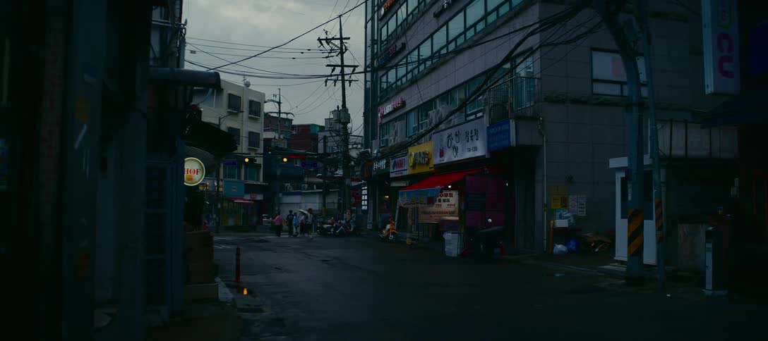 A dark car drives through a gloomy, wet street corner in a South Korean city as pedestrians walk in the background