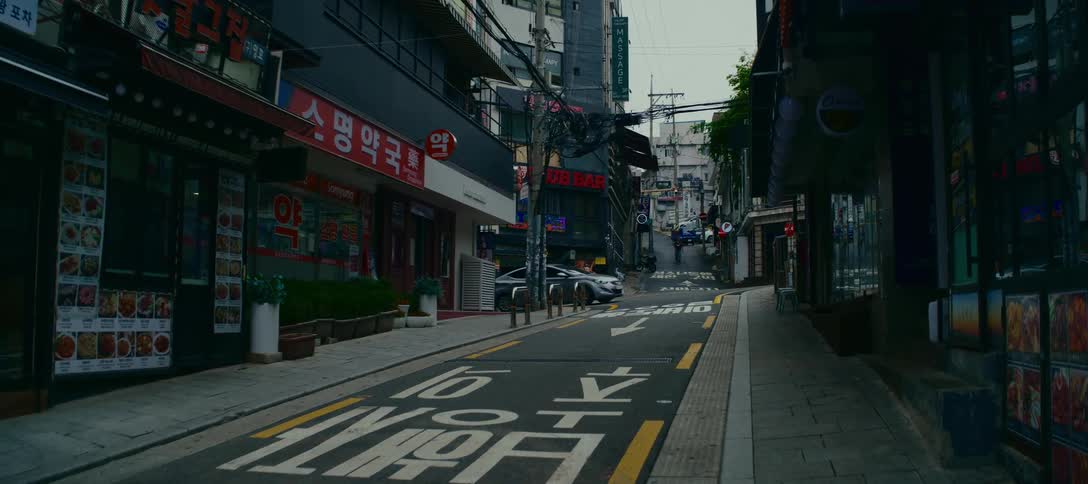 Cars navigate a narrow, hilly backstreet lined with shops and signs in a quiet Korean city