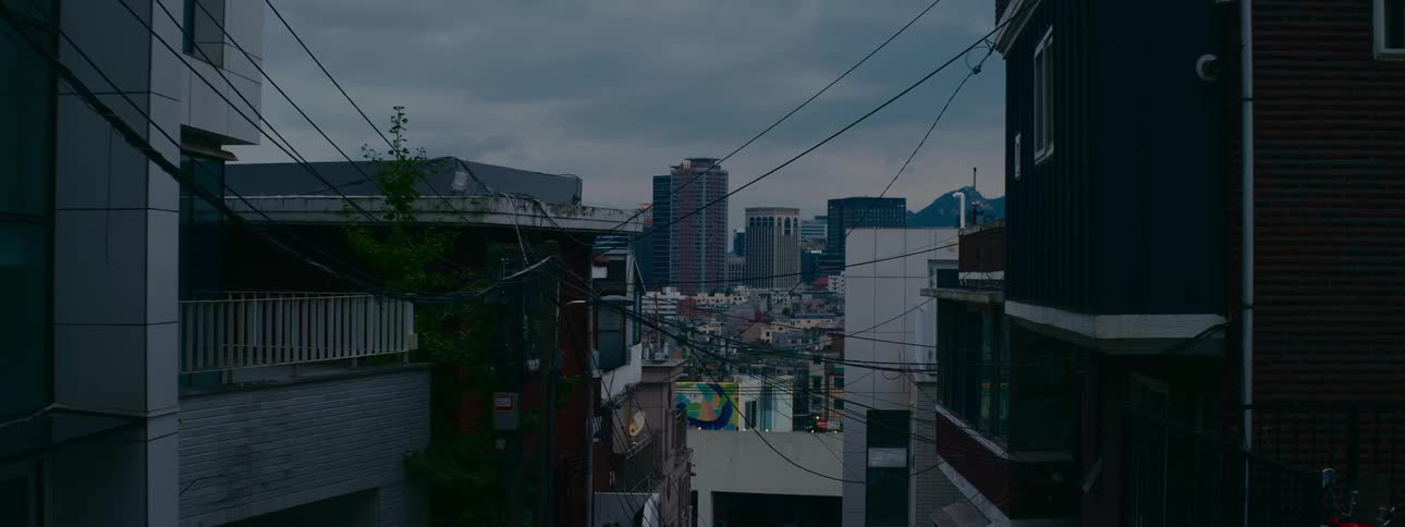 A modern city skyline looms over an older urban neighborhood with tangled power lines on an overcast evening