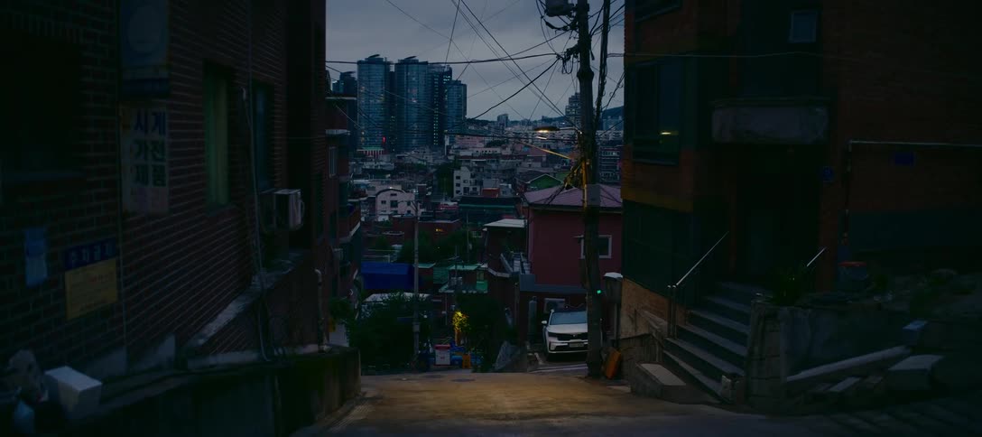 A steep, quiet alleyway in a residential Seoul neighborhood at dusk, with the modern city skyline visible in the background