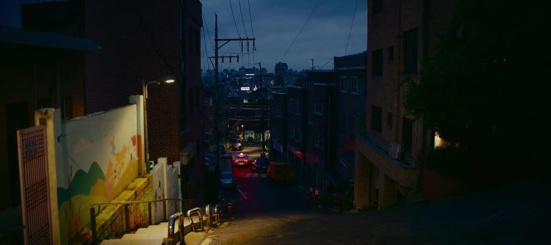 Looking down a steep city street at twilight with distant city lights and a car's red tail lights glowing in the dark