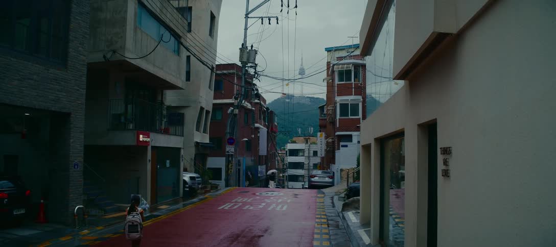A quiet, rainy street in a dense Seoul neighborhood with a view of the distant N Seoul Tower as pedestrians walk by
