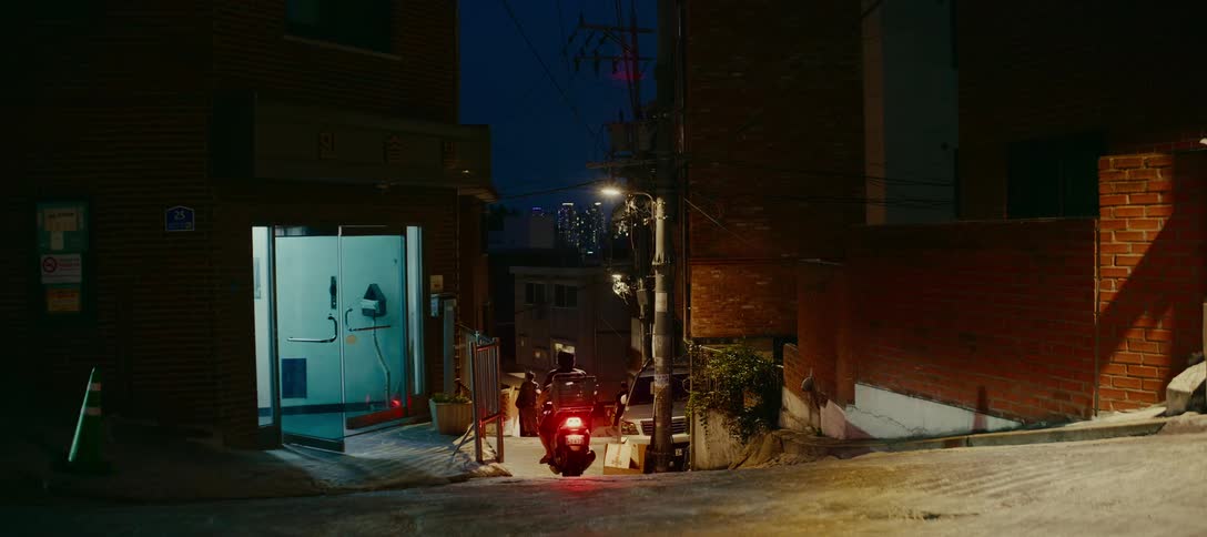 A wide shot captures the nightlife in a steep city alley as delivery drivers on scooters navigate the streets