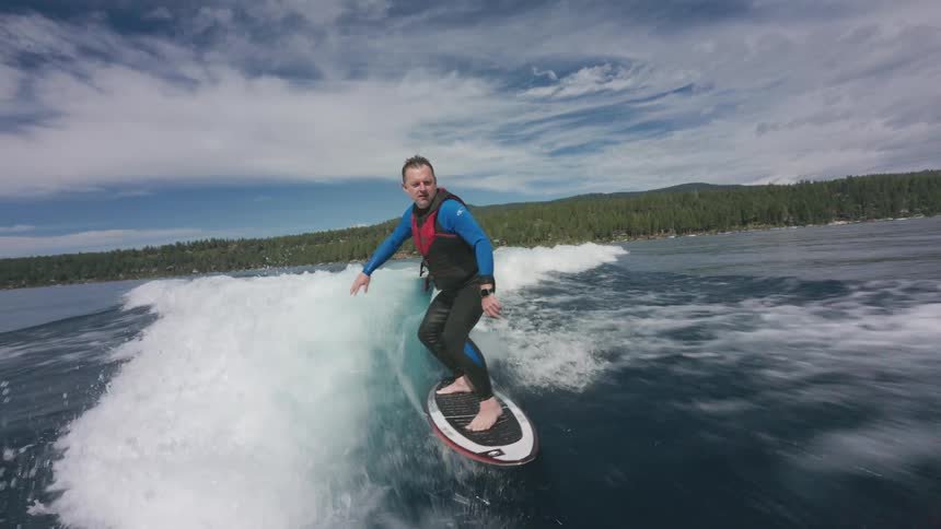 Slow-motion footage follows a man wake surfing on a clear blue lake surrounded by a lush forest