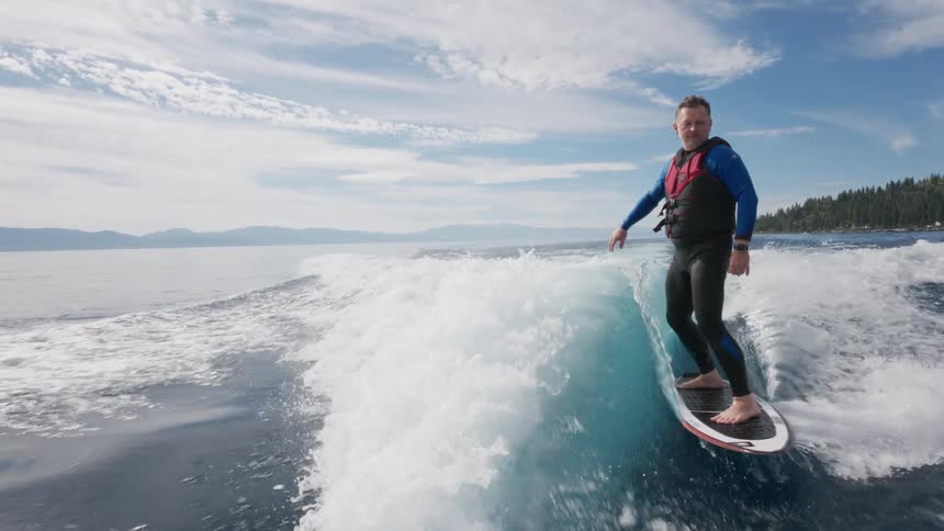 A man in a wetsuit wake surfs in slow motion on a beautiful lake under a partly cloudy sky