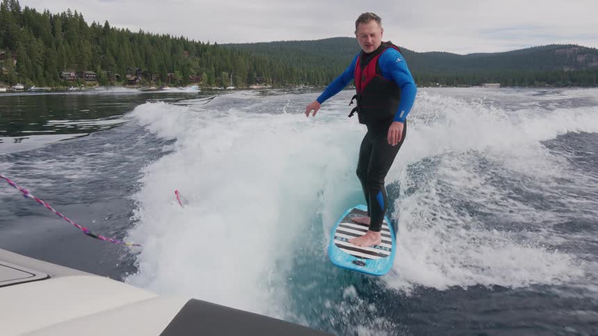 A man in a wetsuit and life vest wakesurfs behind a boat in slow motion, riding the wake on a scenic lake surrounded by forests