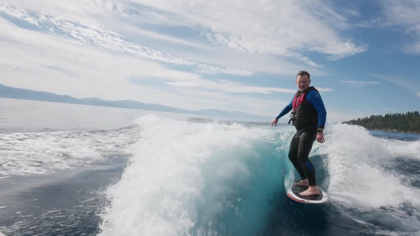 A man in a wetsuit gracefully wakesurfs in slow motion on a beautiful sunny day on a clear blue lake
