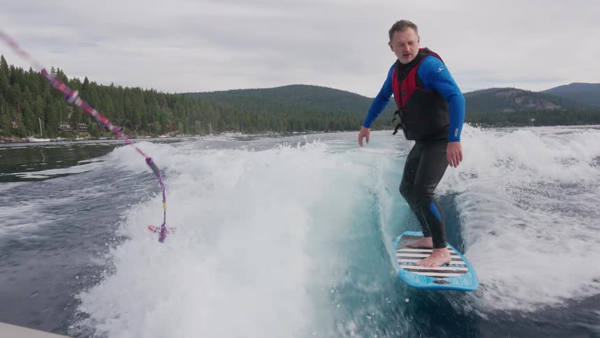 Slow motion shot of a man in a wetsuit wakesurfing behind a boat on a beautiful mountain lake