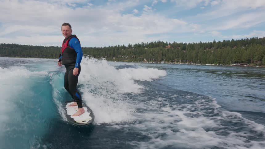 A smiling man in a wetsuit gives a thumbs up while wakesurfing on a clear blue lake in slow motion