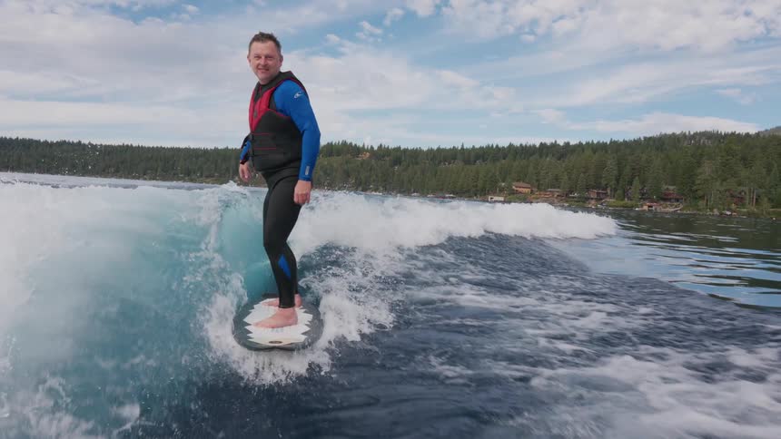 A smiling man in a wetsuit enjoys wakesurfing on a beautiful lake in slow motion