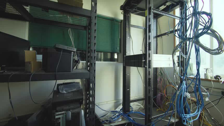 A messy and disorganized server room with a tangled mess of cables on a rack and old computer equipment on shelves