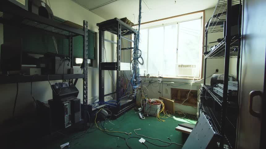 A slow pull-back revealing a messy and neglected server room full of old computer equipment and a chaotic mess of tangled wires