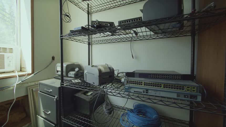 A dilapidated office server room with messy cables and outdated network switches on a wire rack