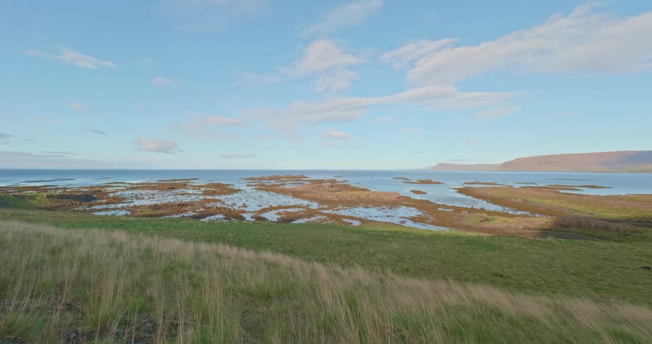 A scenic, wide shot from a grassy hill reveals a tranquil bay with rocky islets in the Icelandic countryside