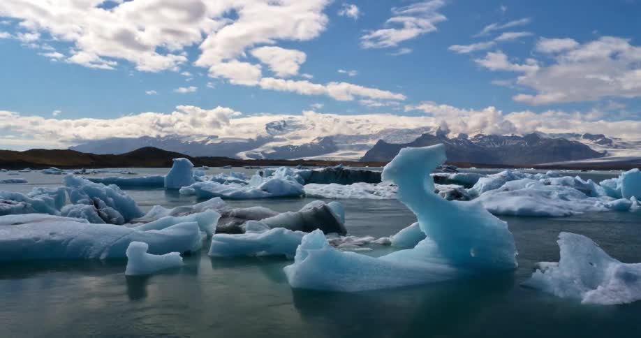 Timelapse of clouds drifting over a tranquil glacial lagoon filled with blue icebergs and distant snowy mountains in Iceland