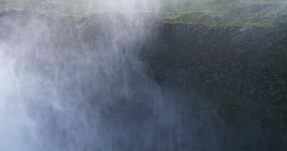 A powerful close-up shot shows a waterfall crashing down a moss-covered cliff in mesmerizing slow motion, creating a thick wall of mist