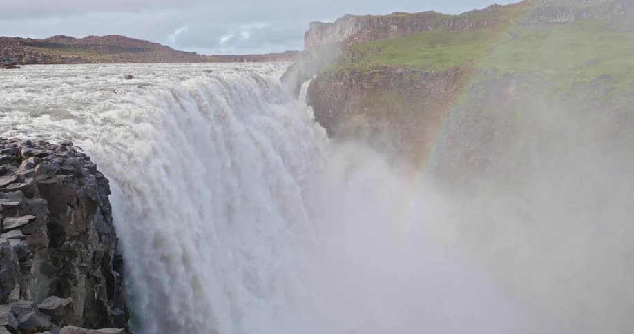 A powerful waterfall thunders over a rocky cliff into a deep canyon, with a faint rainbow appearing in the heavy mist