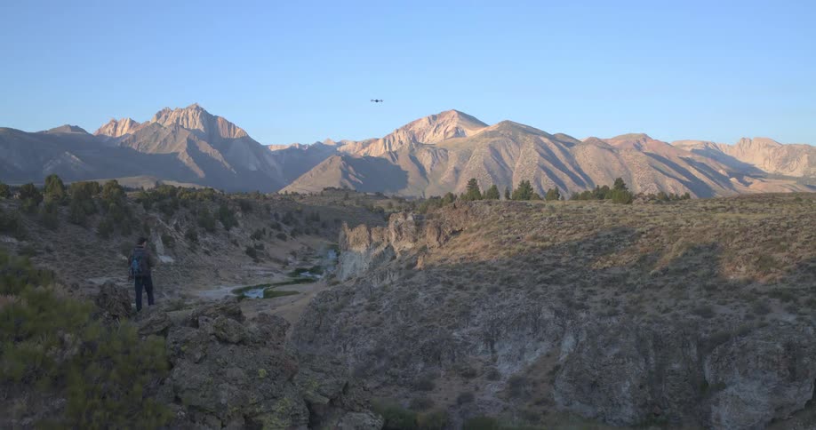 A hiker stands on a cliff's edge flying a drone over a stunning mountain canyon during a golden sunrise