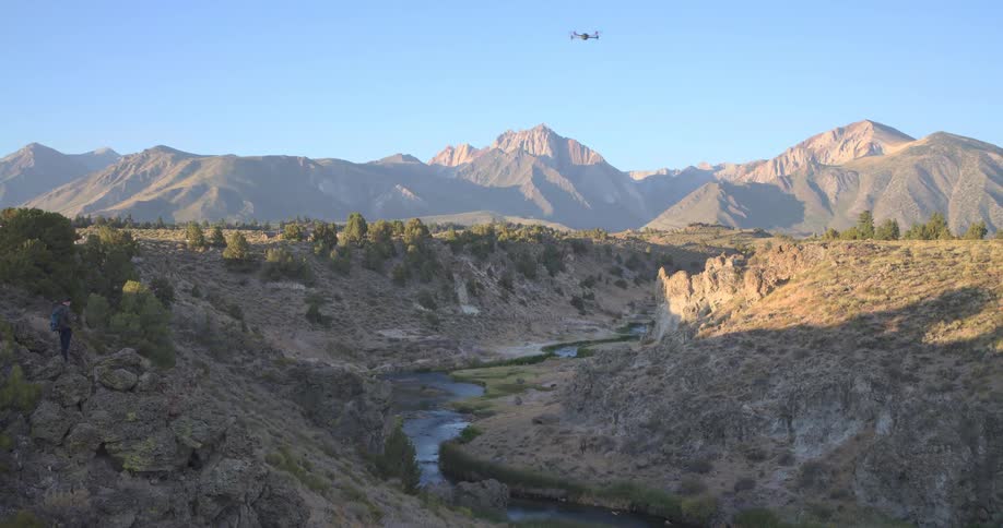 A drone flies over a majestic mountain river valley as its operator stands on a rocky outcrop during golden hour