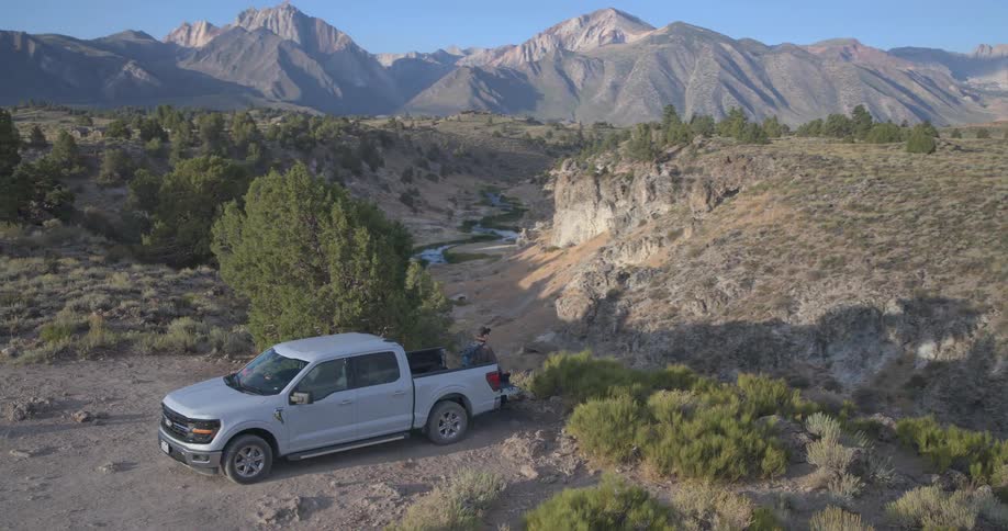 An adventurer sits on the tailgate of his truck, enjoying the scenic view of a river canyon and majestic mountain range