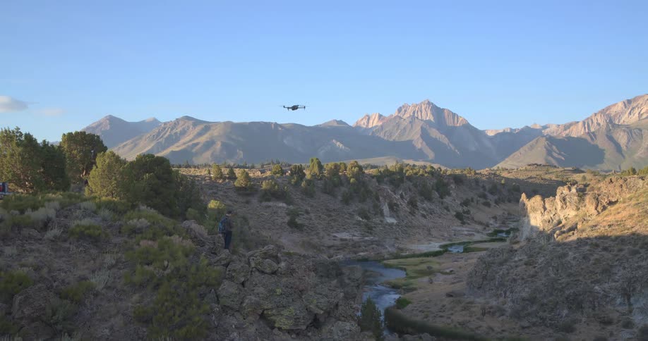 A man pilots a drone through a scenic river canyon with a majestic mountain range in the background