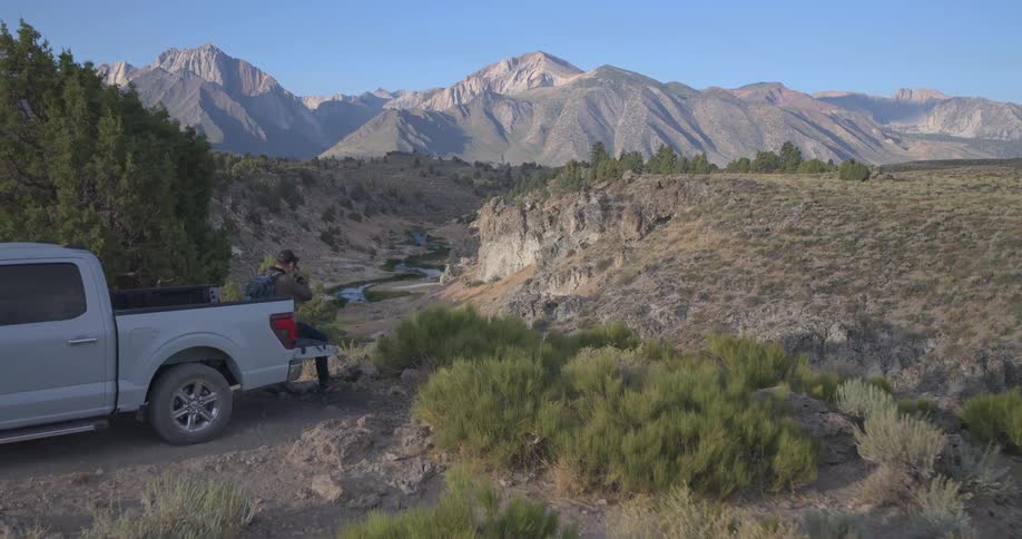 A photographer sits on the tailgate of his truck, capturing the scenic mountain valley and river below as the camera pushes past him