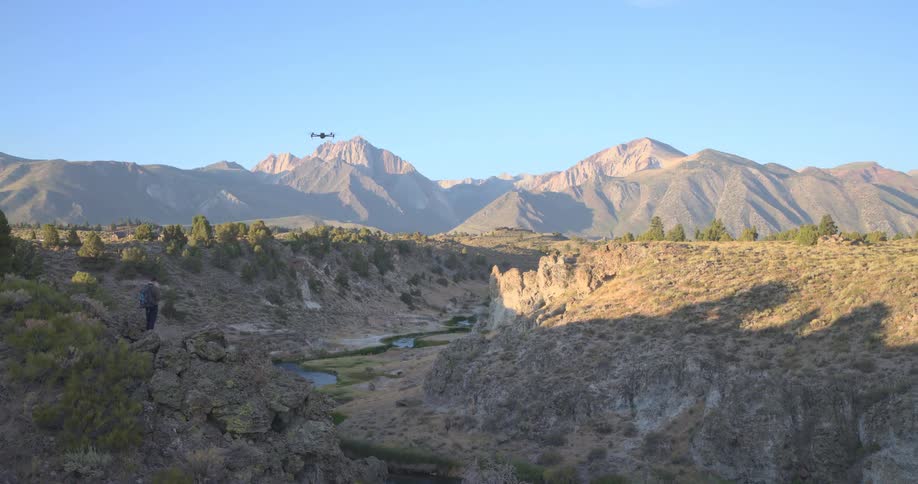 A man pilots a drone in slow motion over a river canyon with a stunning mountain range in the background