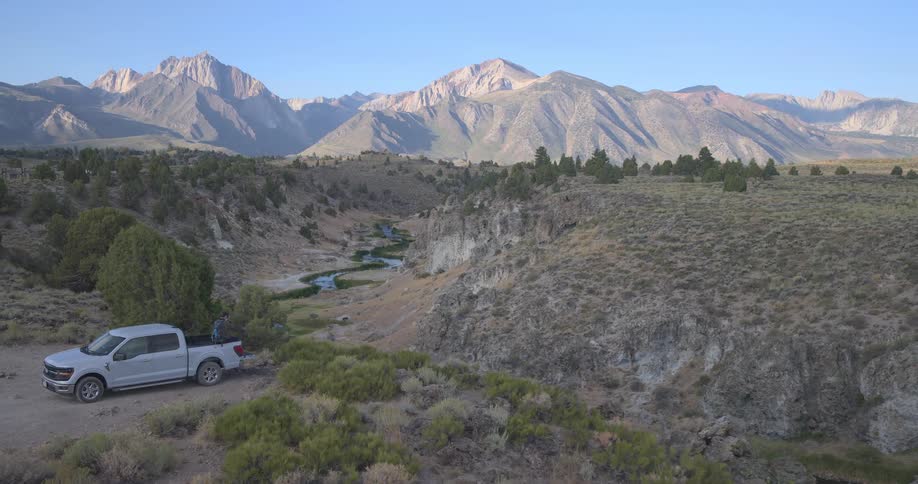 An aerial shot slowly pushes towards a man sitting on the tailgate of a pickup truck, taking in the vast mountain landscape and river canyon below