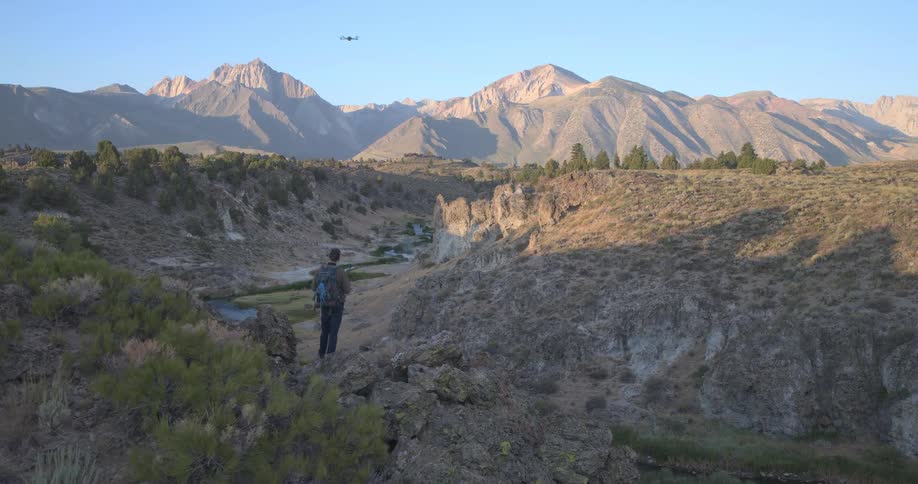 A hiker flies a drone over a stunning river canyon with the majestic Eastern Sierra mountains in the background at sunrise