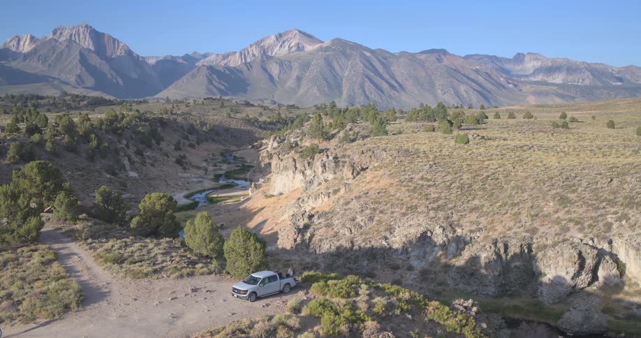An aerial view pulls back to reveal a majestic mountain landscape with a river winding through a canyon and a pickup truck parked on the edge