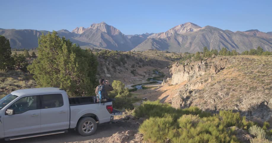 A traveler in a pickup truck takes a break on an overlook, admiring the breathtaking vista of a river winding through a valley with the Sierra Nevada mountains in the background