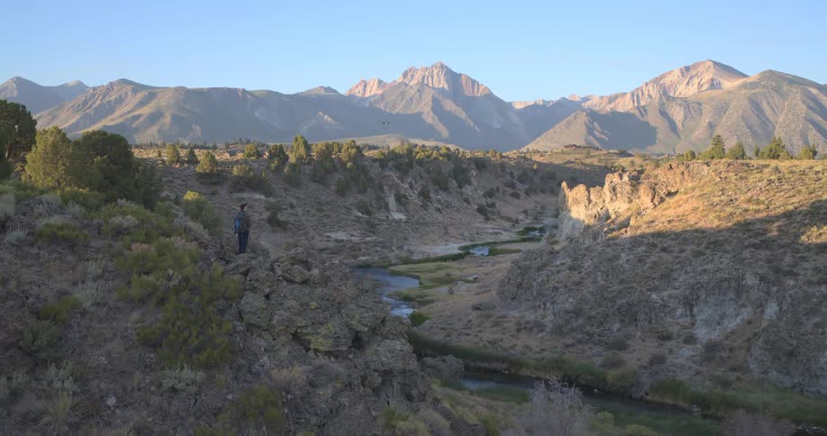 A hiker with a backpack stands on a rocky overlook, admiring the stunning view of a winding river and majestic mountain range during sunrise
